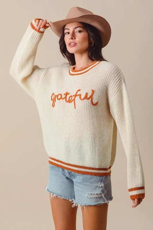 Woman wearing a cream sweater with 'grateful' text, denim shorts, and a beige hat on a beige background