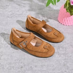 A pair of tan suede Mary Jane style shoes with buckle straps, displayed on a gray textured floor next to a pink flower pot.