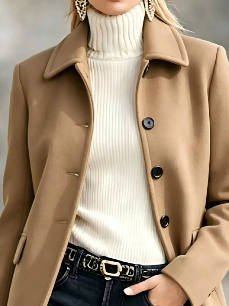 A close-up of a person wearing a camel brown coat over a cream ribbed turtleneck sweater, paired with dark jeans and a black belt with a silver buckle.