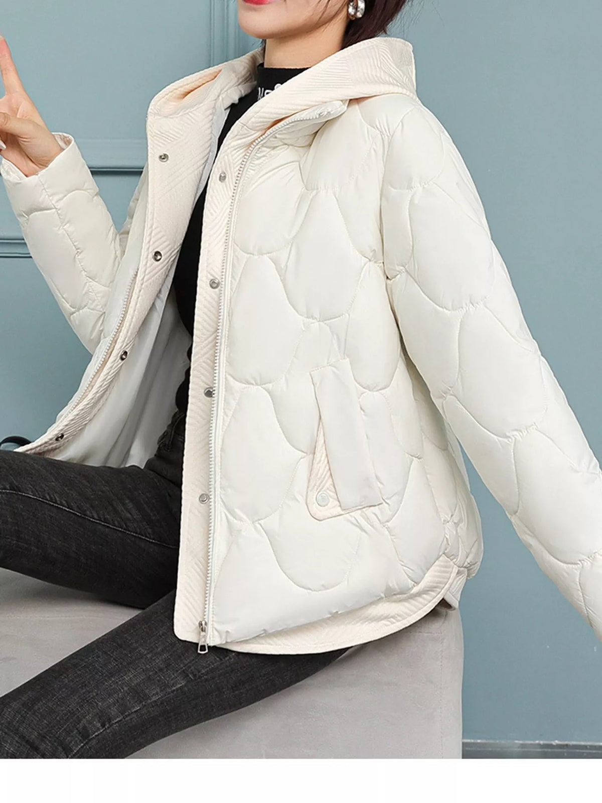 A woman wearing a cream-colored quilted puffer jacket with a hood, layered over a black top and dark jeans, seated against a light blue-gray paneled wall.