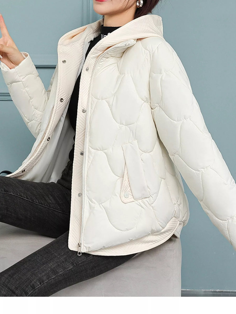 A woman wearing a cream-colored quilted puffer jacket with a hood, layered over a black top and dark jeans, seated against a light blue-gray paneled wall.