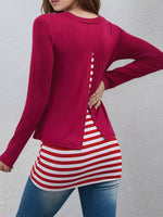 A woman wearing a long-sleeve magenta top with a back slit detail, layered over a red and white striped t-shirt and blue jeans, shown from the back against a textured gray wall.