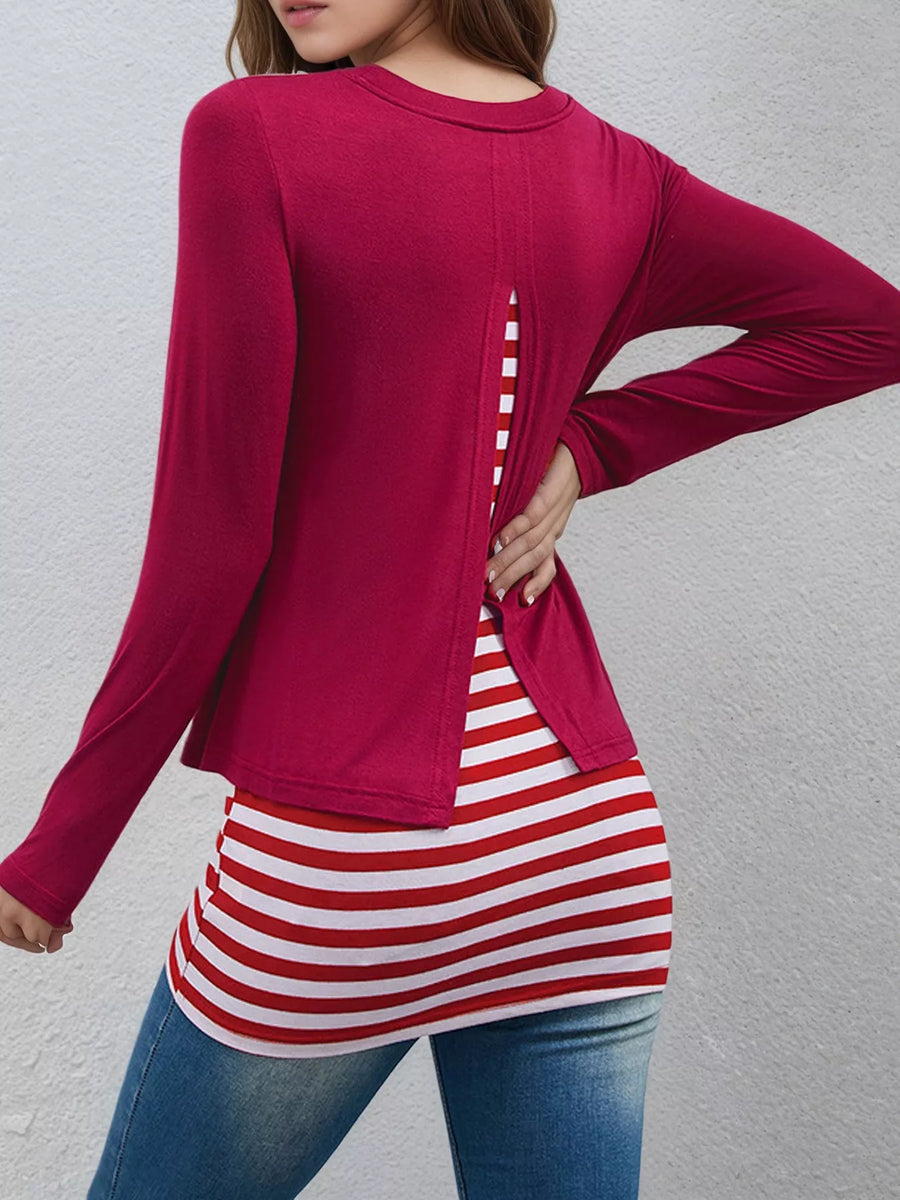 A woman wearing a long-sleeve magenta top with a back slit detail, layered over a red and white striped t-shirt and blue jeans, shown from the back against a textured gray wall.