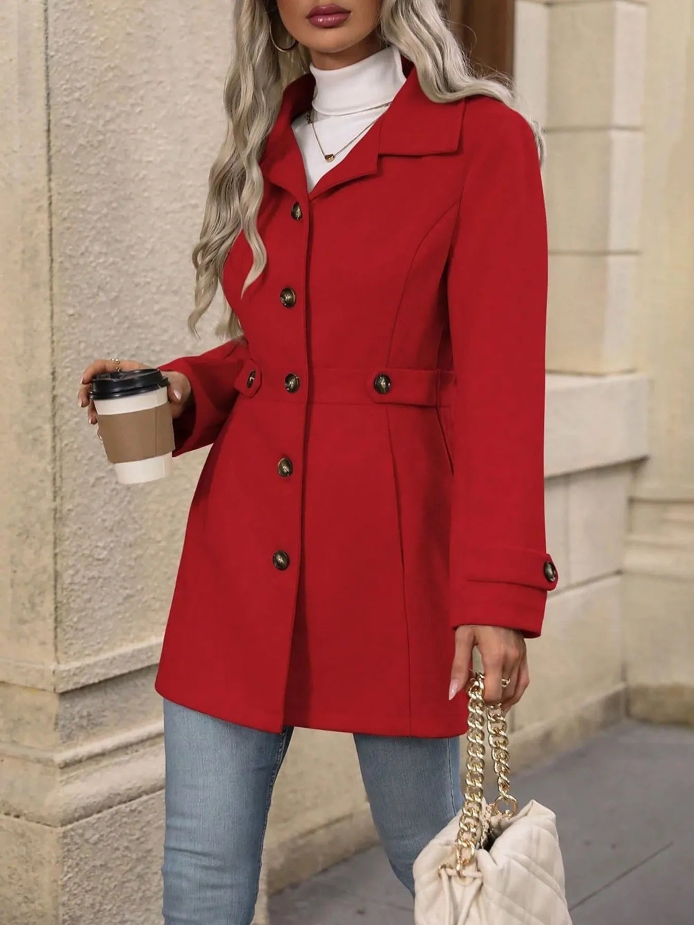 Woman wearing a red coat holding a coffee cup and a handbag on a city street.