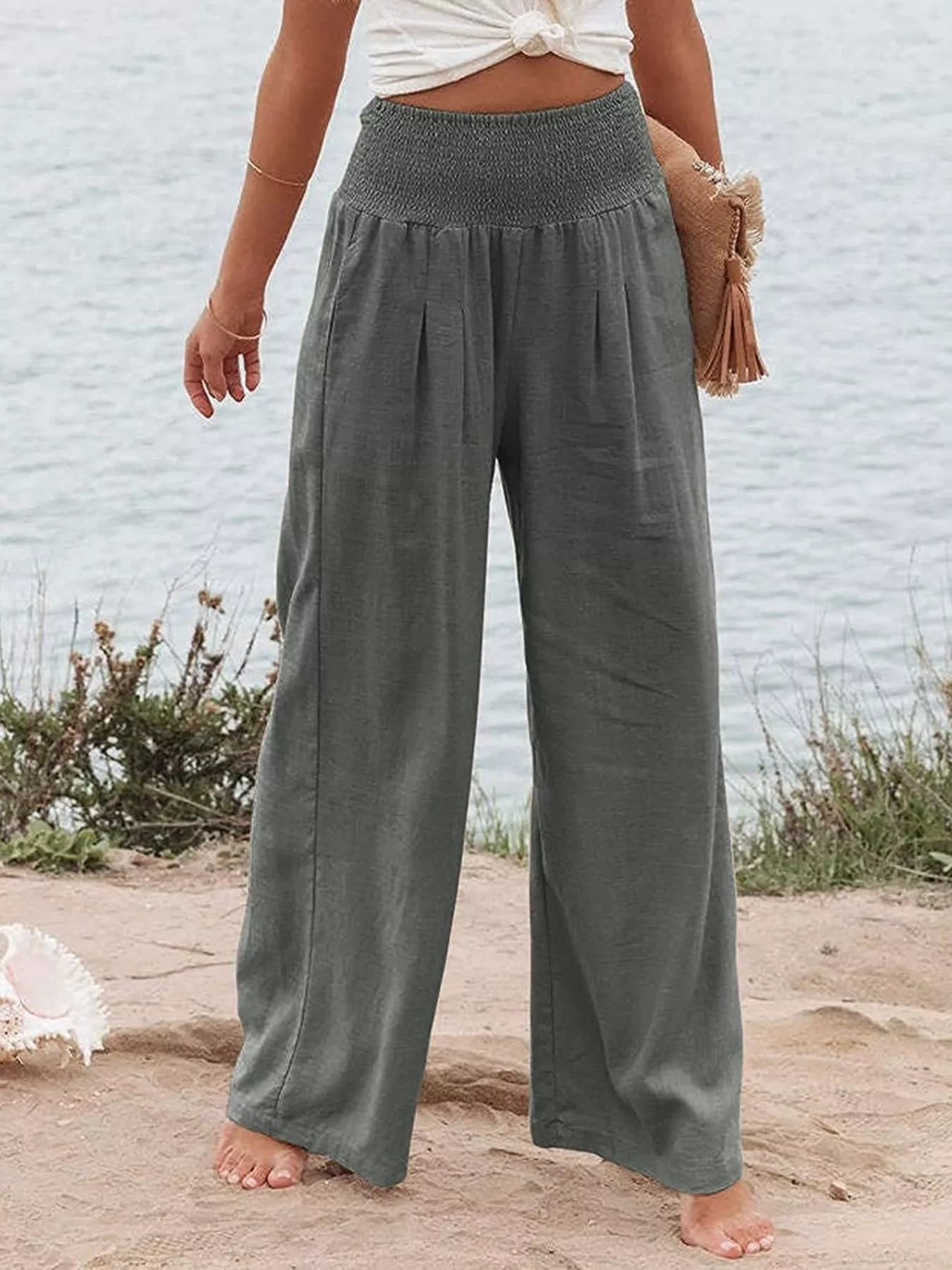Person wearing wide-leg gray pants standing on a sandy beach with a light blue wall in the background.