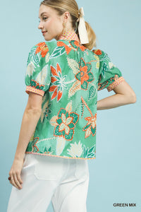 Woman wearing a green floral blouse with white pants against a light blue background