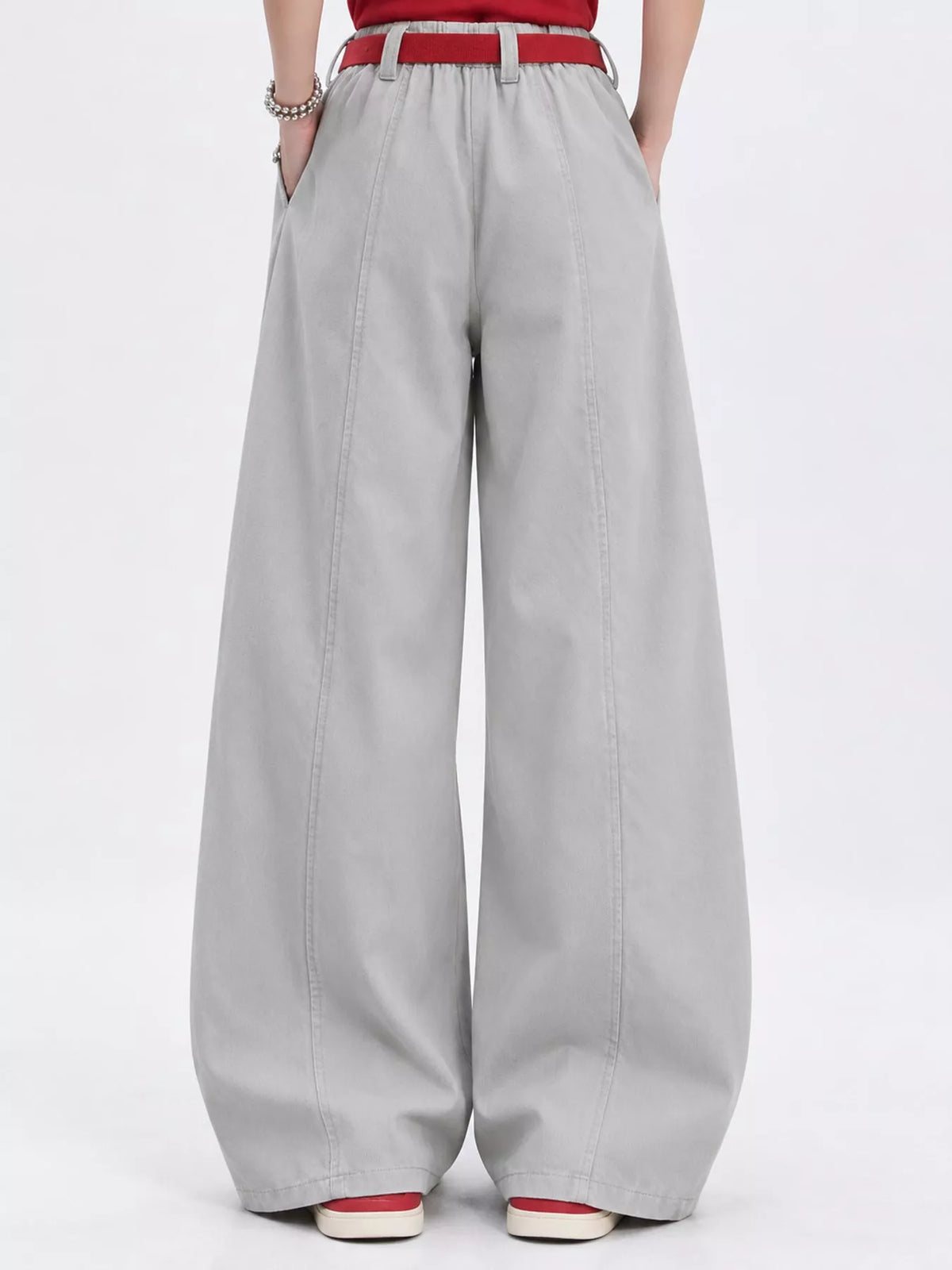 A person shown from the waist down wearing light gray wide-leg trousers with a red belt, a red top, and red-and-white shoes against a plain white studio background.