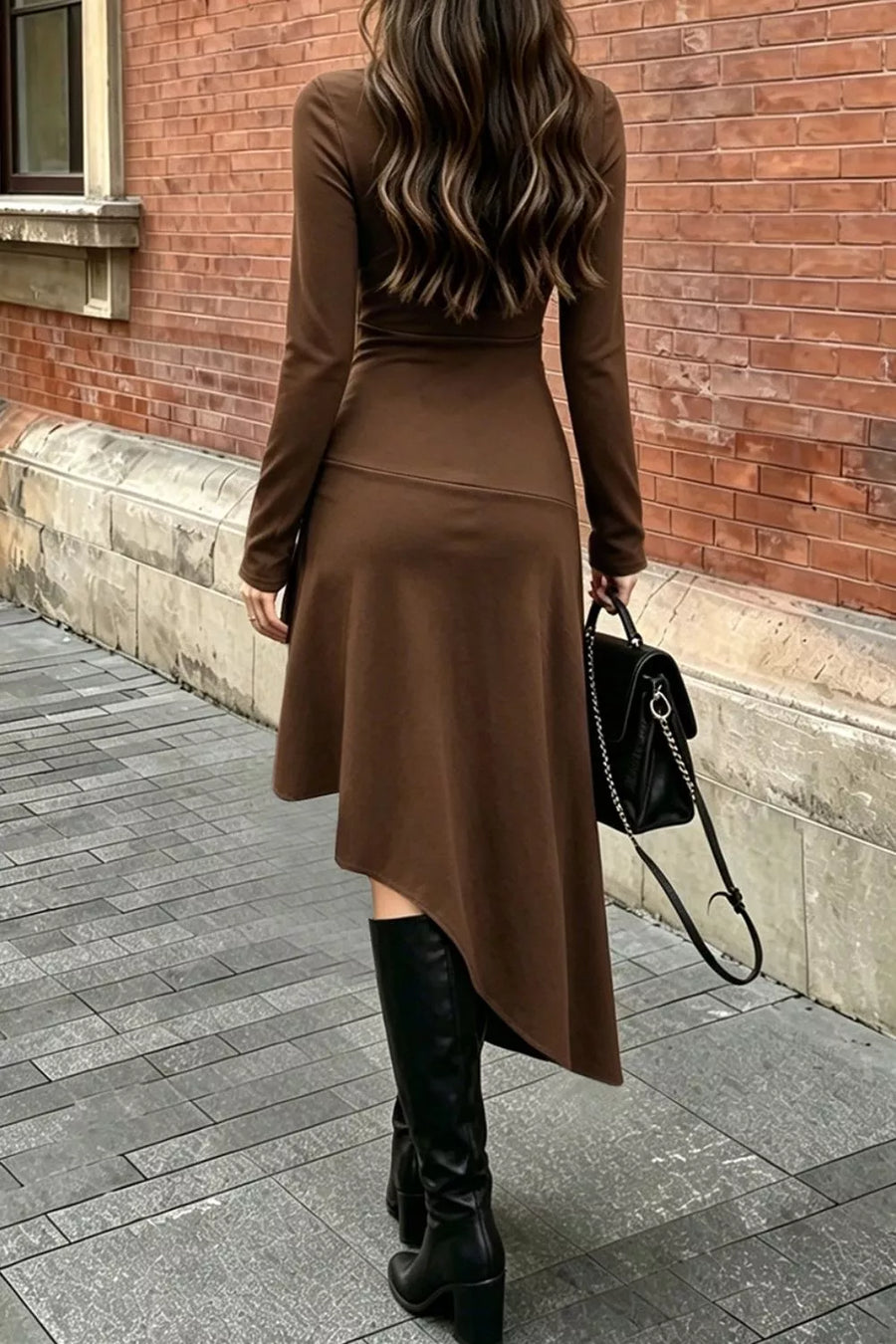 A woman viewed from behind wearing a fitted long-sleeve brown midi dress with an asymmetrical hem, paired with black knee-high boots and carrying a black handbag, standing on a paved sidewalk against a red brick wall.
