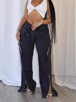 A woman modeling black wide-leg cargo pants with silver zippers and side pockets, paired with a white cutout top, standing in front of a white curtain backdrop.