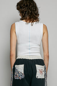 Person wearing a white sleeveless top and black pants with floral pockets on a gray background