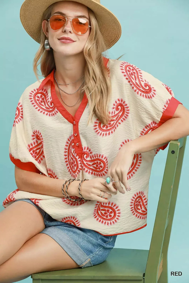 Woman wearing a red and white patterned shirt with a straw hat and sunglasses, sitting on a green chair against a light blue background.