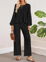 Woman wearing a black outfit with wide-leg pants, holding a brown bag against a white wall with green leaves.