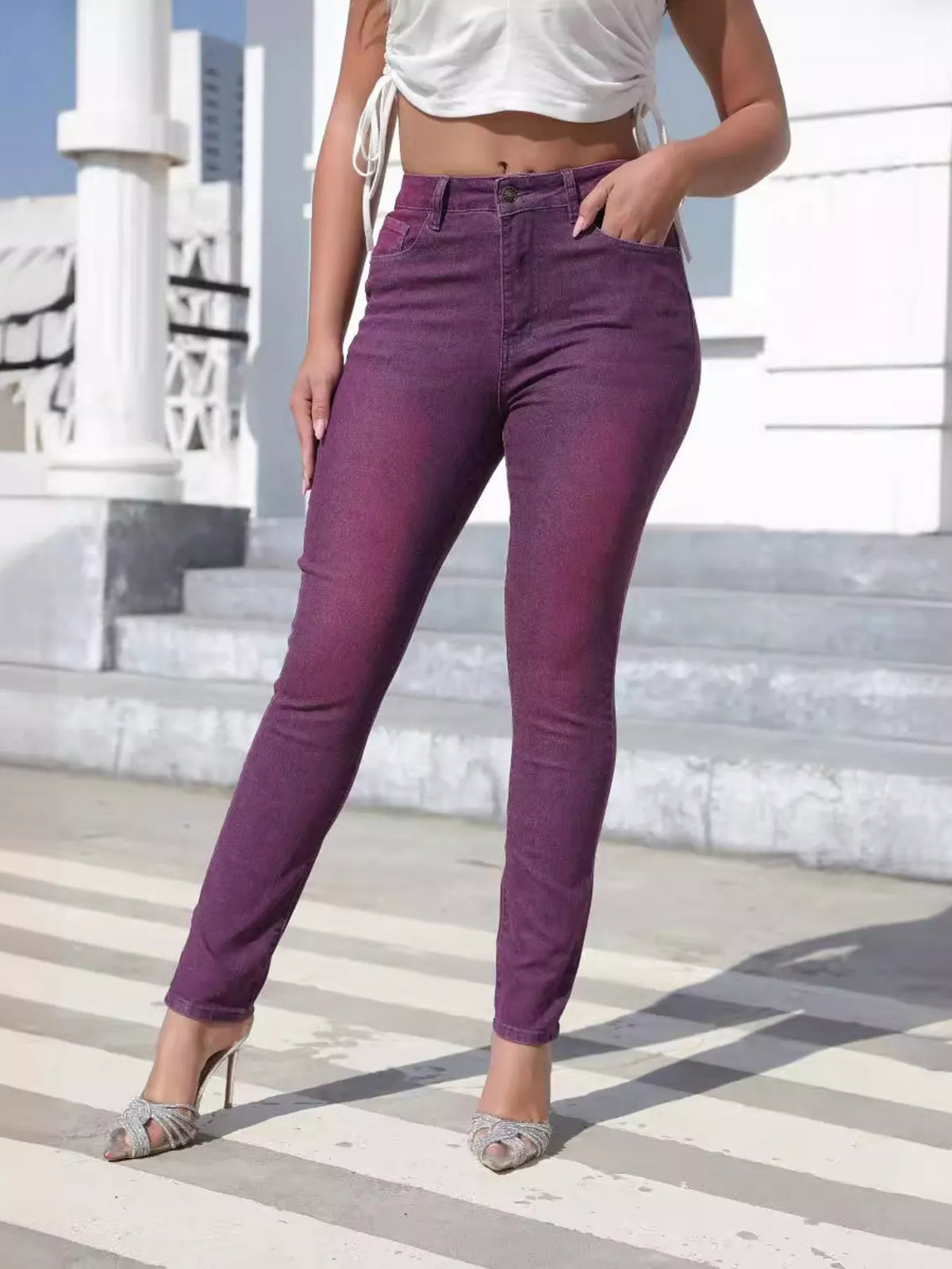 A model wearing high-waisted, skinny purple jeans with a faded wash, styled with a white crop top and silver heels, standing on outdoor concrete steps.