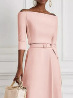 Woman wearing a pink off-shoulder dress with a belt, holding a woven bag against a white background.