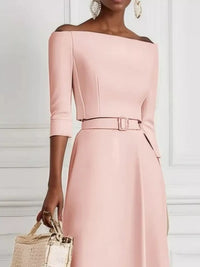 Woman wearing a pink off-shoulder dress with a belt, holding a woven bag against a white background.