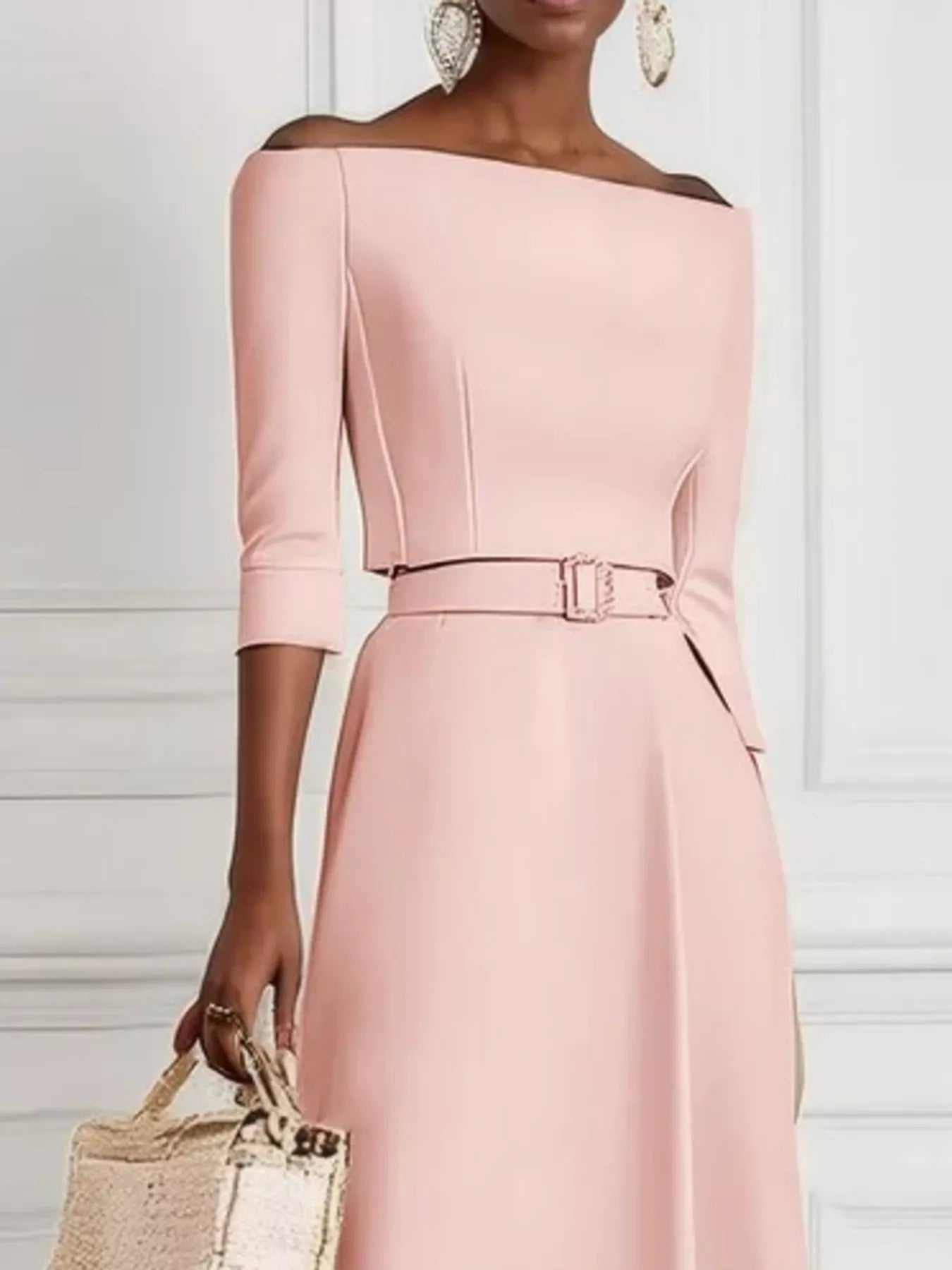 Woman wearing a pink off-shoulder dress with a belt, holding a woven bag against a white background.