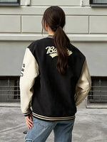 A woman viewed from behind wearing a black and cream varsity jacket with gold lettering on the back, paired with blue jeans, standing outdoors against a light gray brick wall.