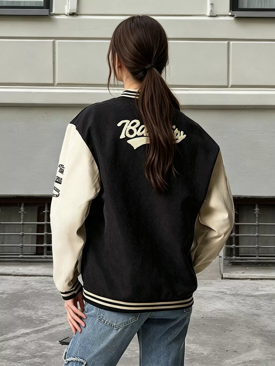 A woman viewed from behind wearing a black and cream varsity jacket with gold lettering on the back, paired with blue jeans, standing outdoors against a light gray brick wall.