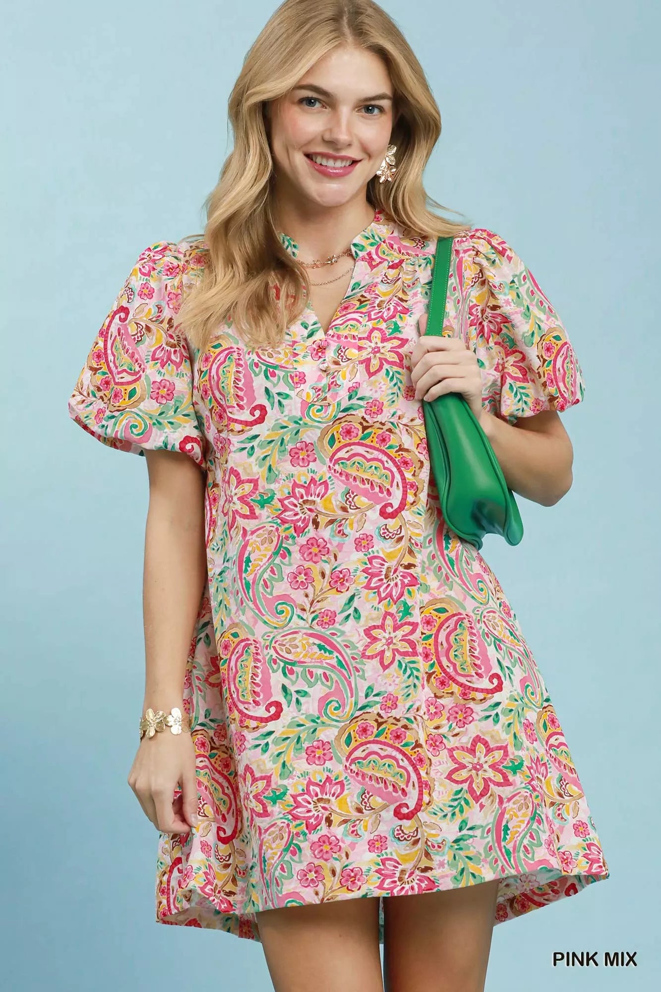 Woman wearing a colorful patterned dress holding a green bag against a light blue background