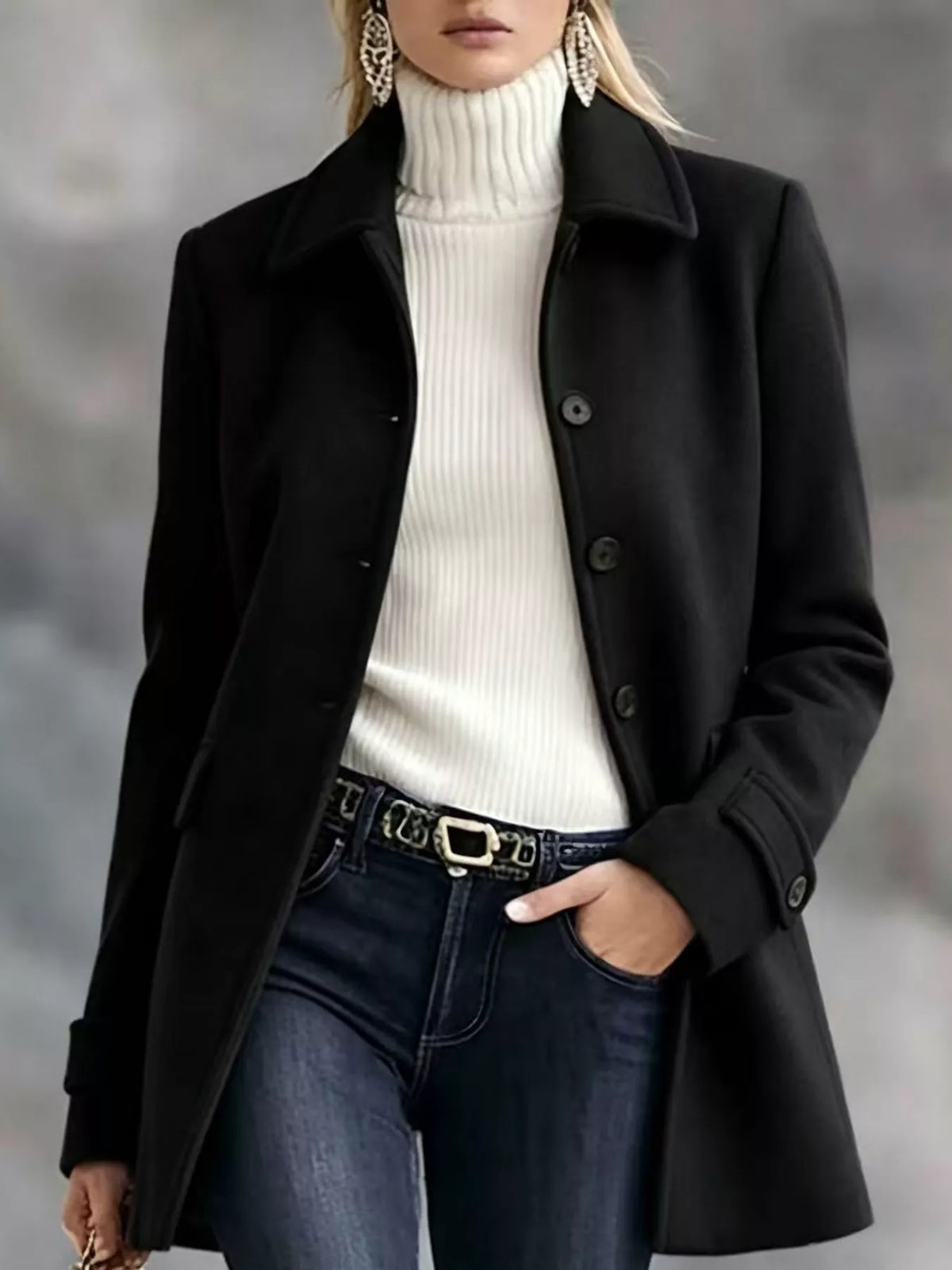 A woman wearing a black mid-length coat over a white ribbed turtleneck sweater and dark blue jeans, with one hand in a pocket and the other holding a phone. She is wearing large statement earrings.