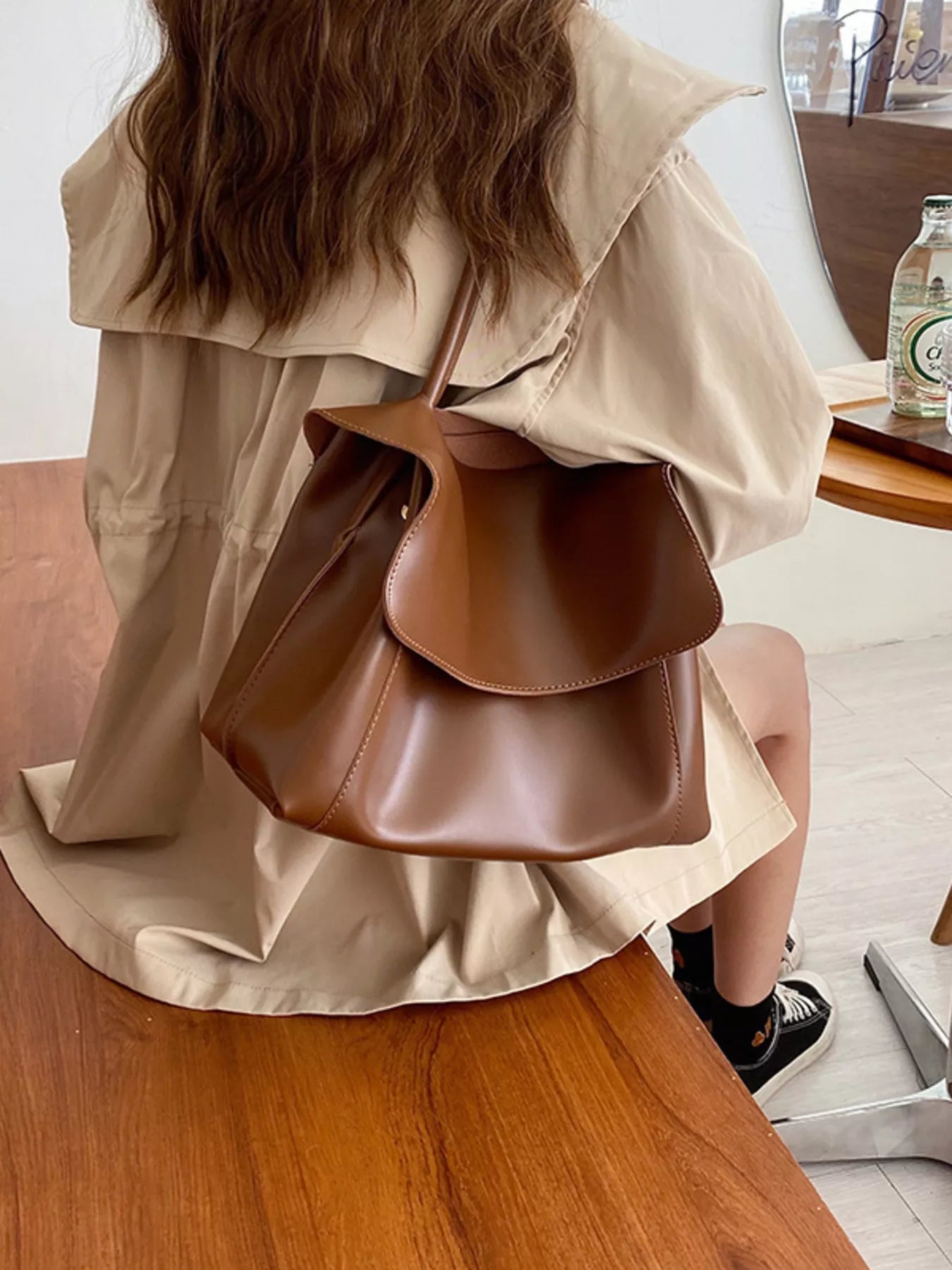 Person wearing a beige trench coat holding a brown leather bag indoors.
