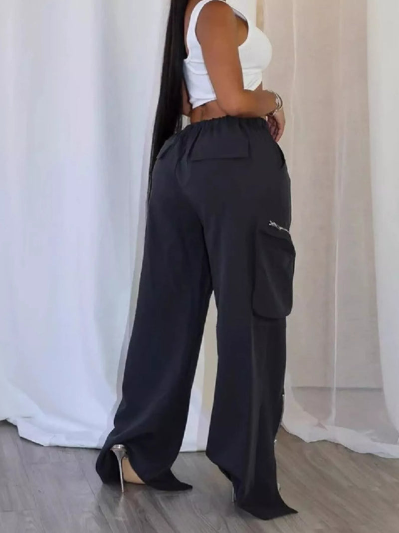 A woman wearing black wide-leg cargo pants with side pockets, paired with a white crop top and black pointed-toe heels, standing in front of white draped curtains.