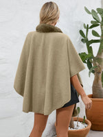 Person wearing a beige cape with fur trim, standing in front of a white brick wall and potted plants.