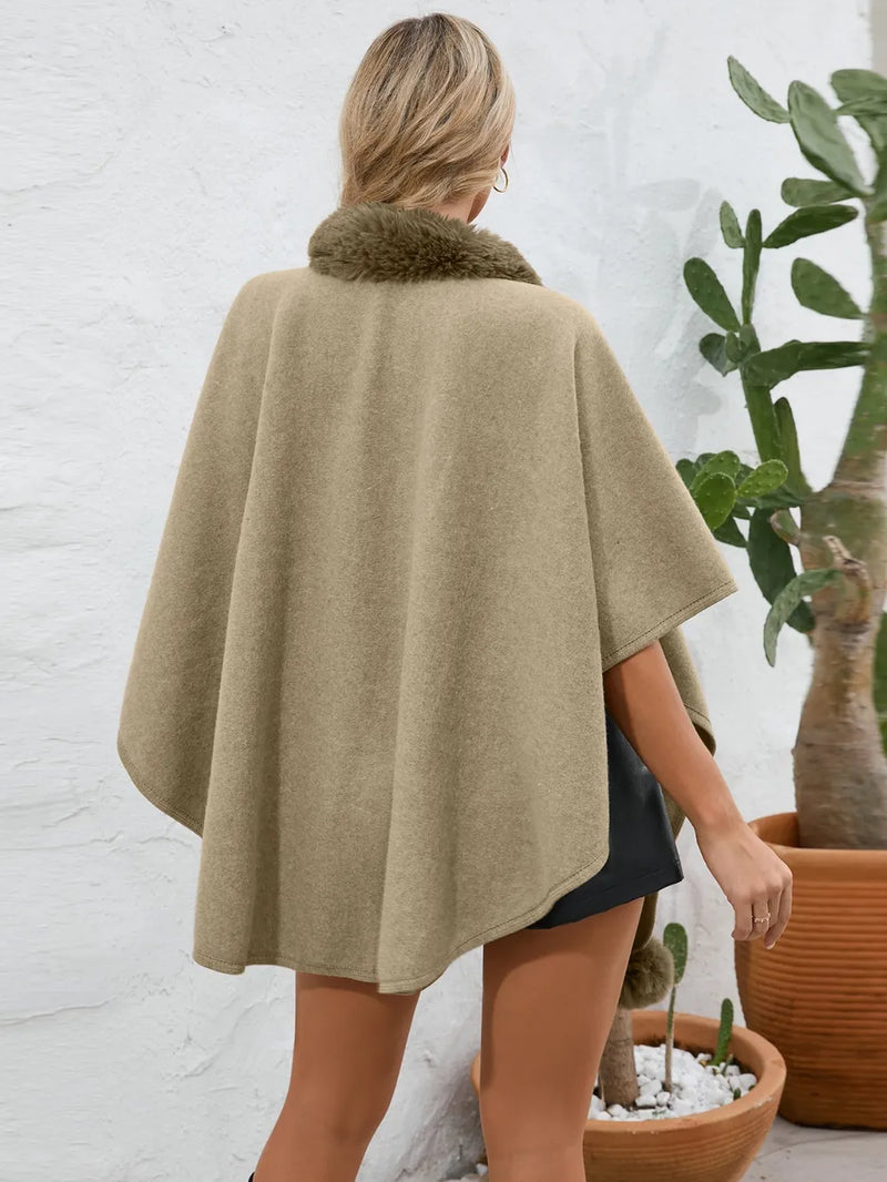 Person wearing a beige cape with fur trim, standing in front of a white brick wall and potted plants.