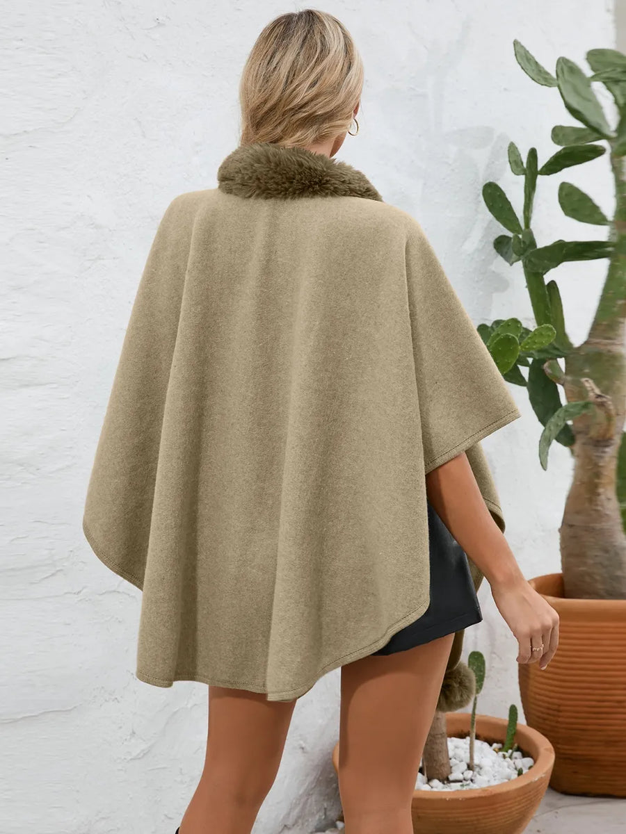 Person wearing a beige cape with fur trim, standing in front of a white brick wall and potted plants.