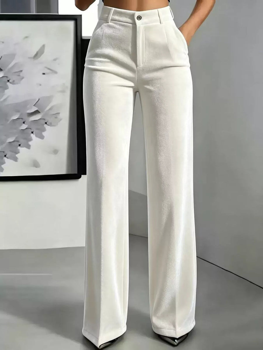 A model wearing a pair of high-waisted, wide-leg white trousers with a subtle textured sheen, paired with black pointed-toe heels, shown from the waist down in an indoor setting.