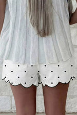 White shorts with black polka dots and scalloped edge worn by a person against a stone wall background.