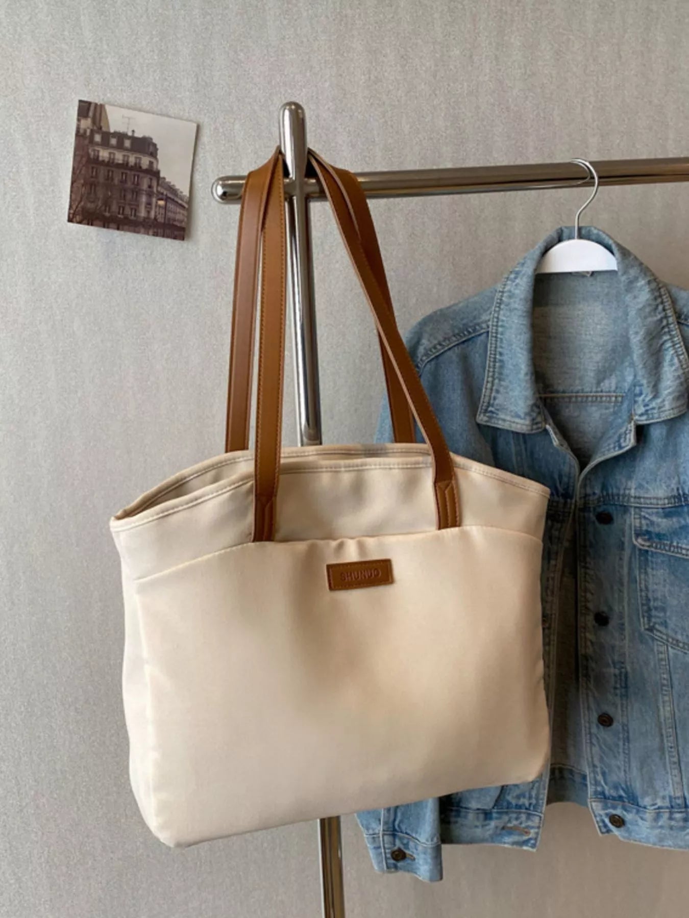 Beige tote bag with brown straps hanging on a coat rack with a denim jacket.