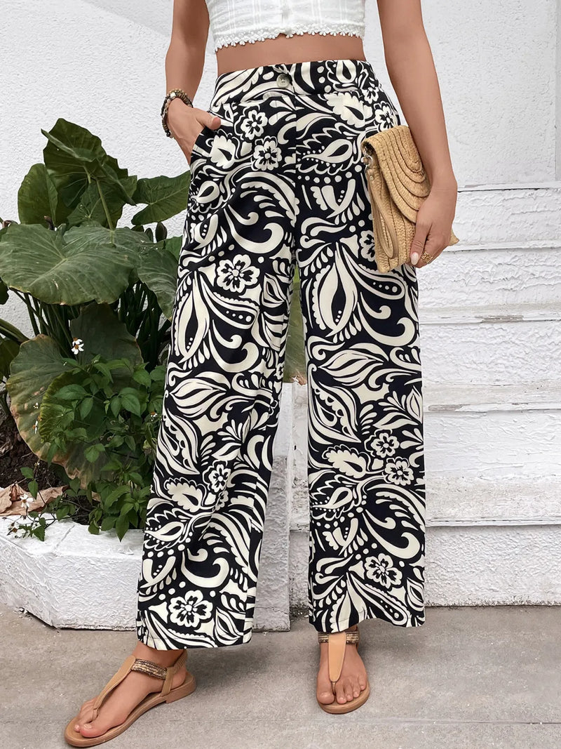 Person wearing black and white patterned pants with a white top and sandals, standing next to a plant.