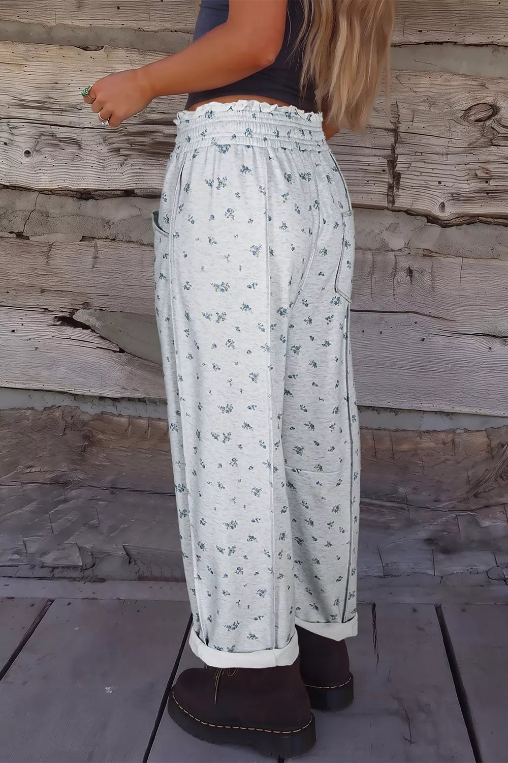 A person shown from the back wearing high-waisted, wide-leg pants with a small blue floral print on a white background, paired with dark brown boots, standing against a rustic wooden wall.