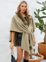 Woman wearing a beige shawl with fur trim in front of a white wall and potted plants.
