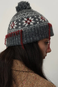 Person wearing a gray knit hat with red accents and a pom-pom on a plain background