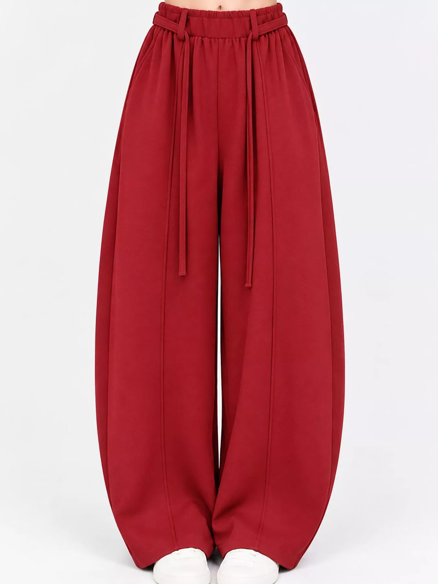 A pair of wide-leg, balloon-style pants in a deep red or burgundy color, featuring an elastic waistband with hanging drawstrings, shown on a model from the waist down against a plain white background.