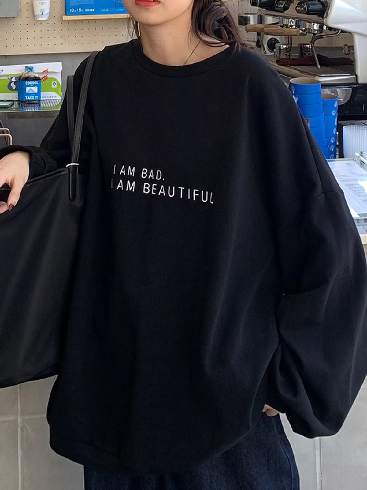 A person wearing an oversized black crewneck sweatshirt with the text 'I AM BAD, I AM BEAUTIFUL' printed in white on the chest, holding a black tote bag in a cafe setting.