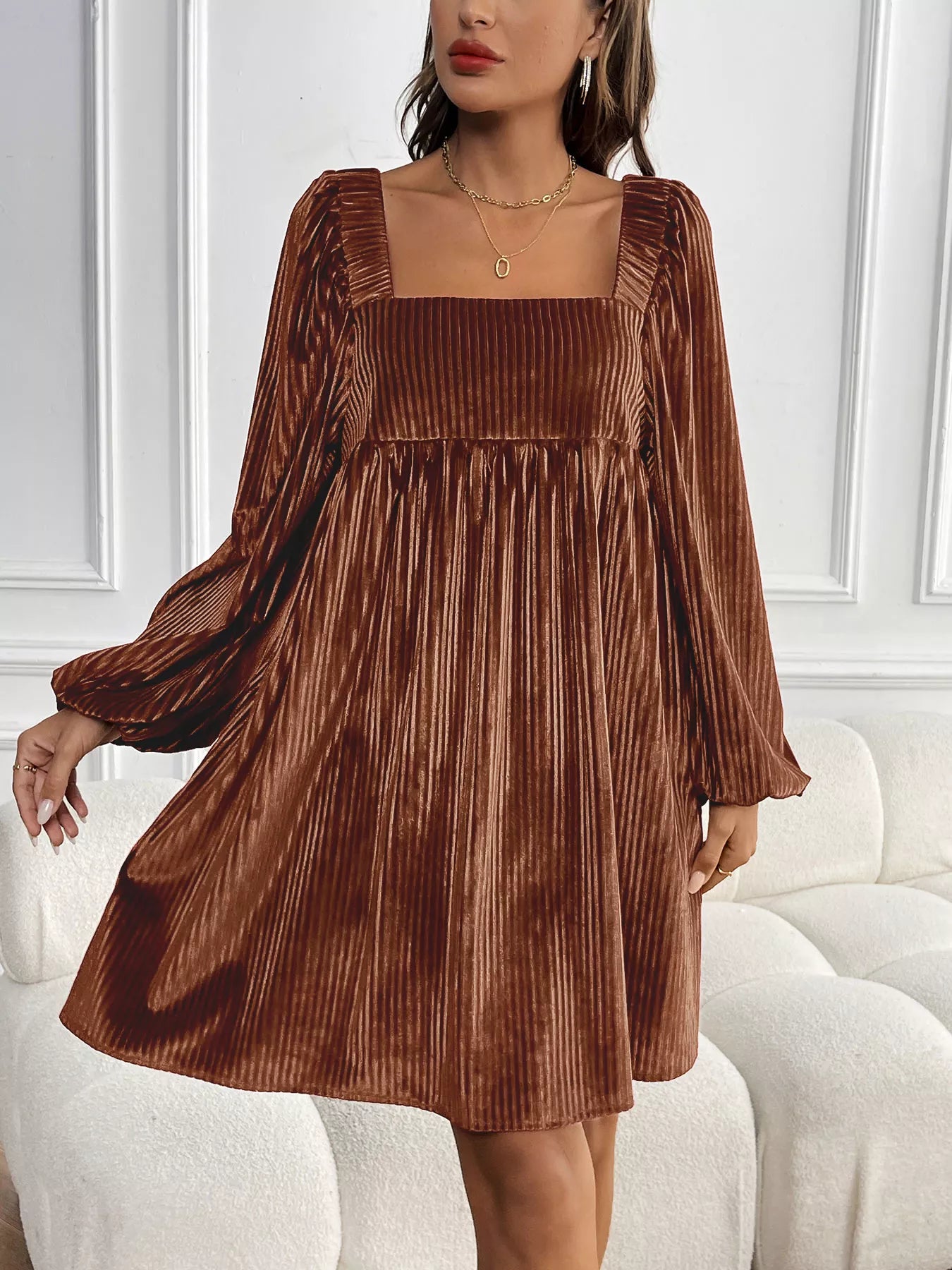 Woman wearing a brown pleated dress against a white wall.