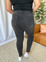 Person wearing black jeans and a black top in an indoor setting with a couch and plant in the background.