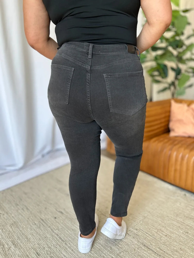 Person wearing black jeans and a black top in an indoor setting with a couch and plant in the background.