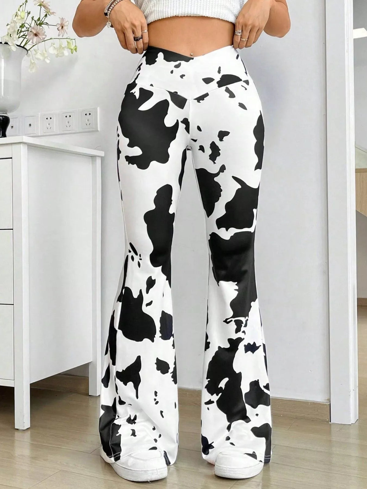 A person wearing high-waisted black and white cow-print flare pants, paired with a white ribbed crop top and white sneakers, standing in a bright indoor room.