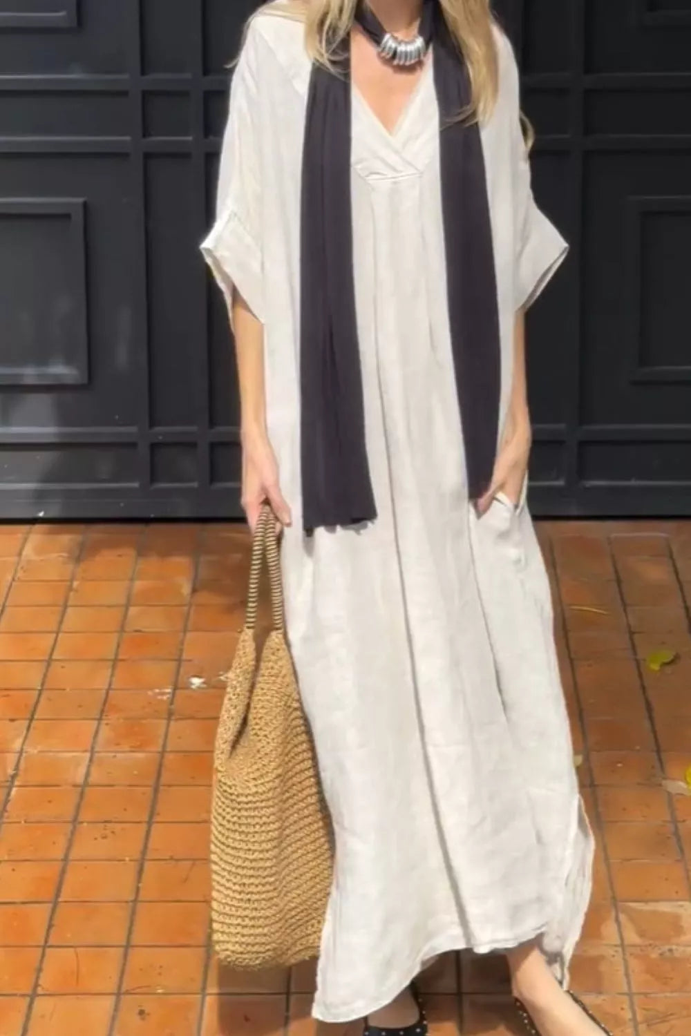 A woman wearing a loose, off-white maxi dress with a dark charcoal scarf, holding a large woven straw tote bag, standing in front of a dark paneled wall and terracotta tiled floor.