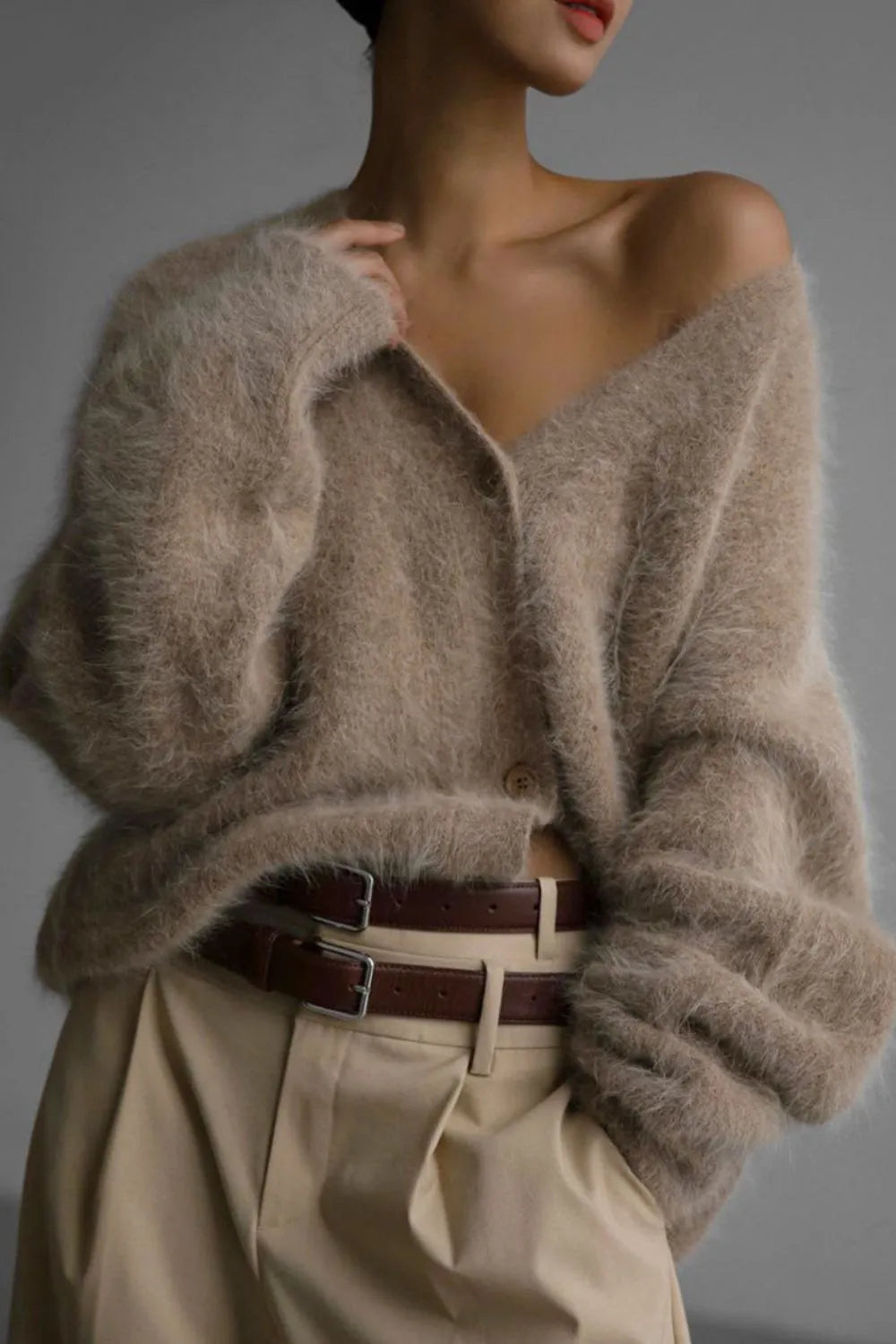 Person wearing a beige cardigan and beige pants with a brown belt against a gray background