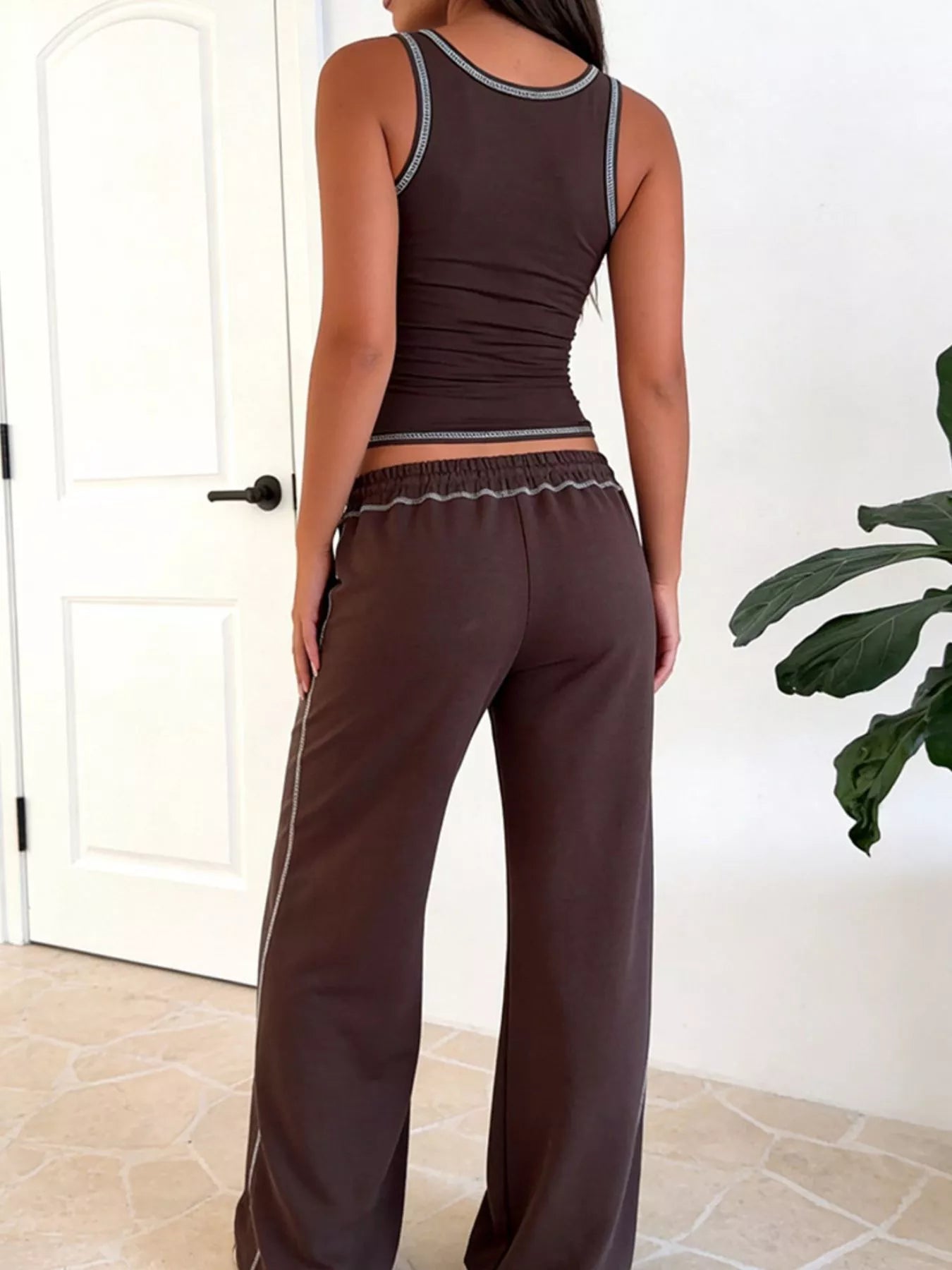 Person wearing a brown tank top and pants in a room with a white door and plant.