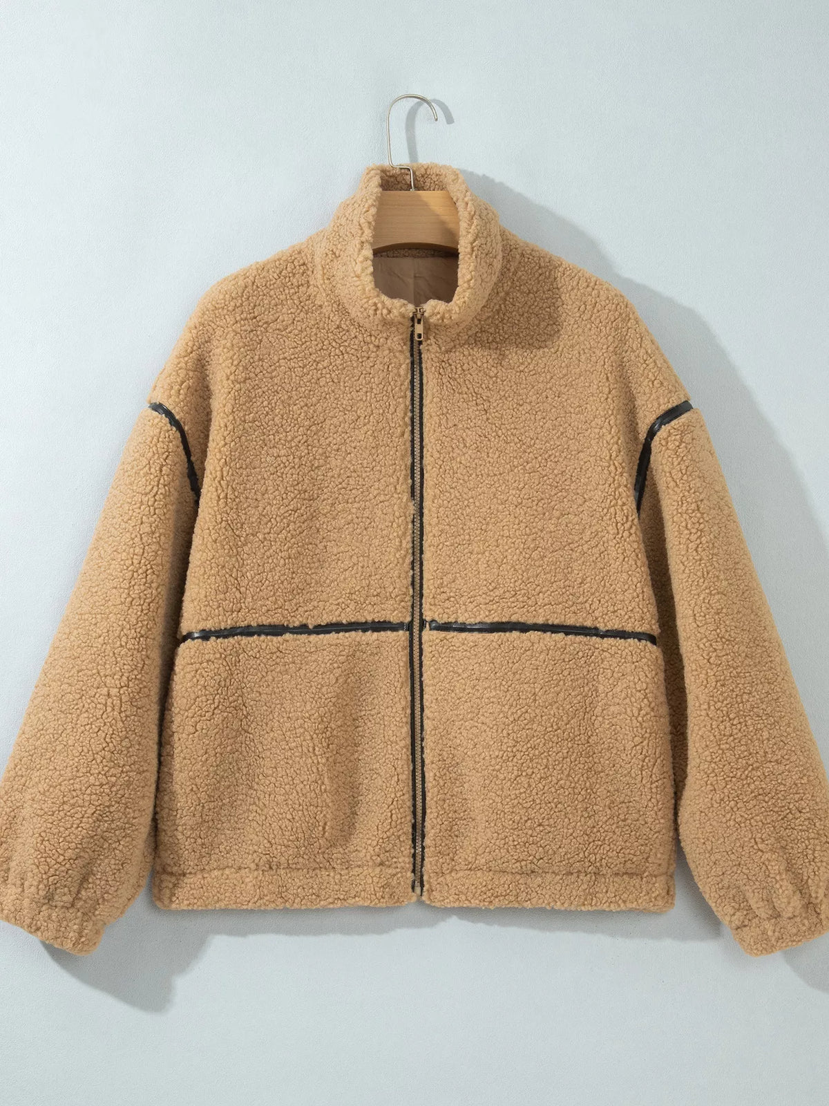 A tan sherpa fleece zip-up jacket with black contrast stitching details, hanging on a wooden hanger against a light gray wall.