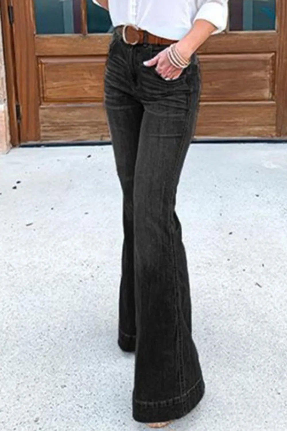 Person wearing black flared jeans standing on a concrete surface with a wooden door in the background.