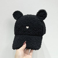 Black knitted bear hat with heart detail held by a hand against a light gray background