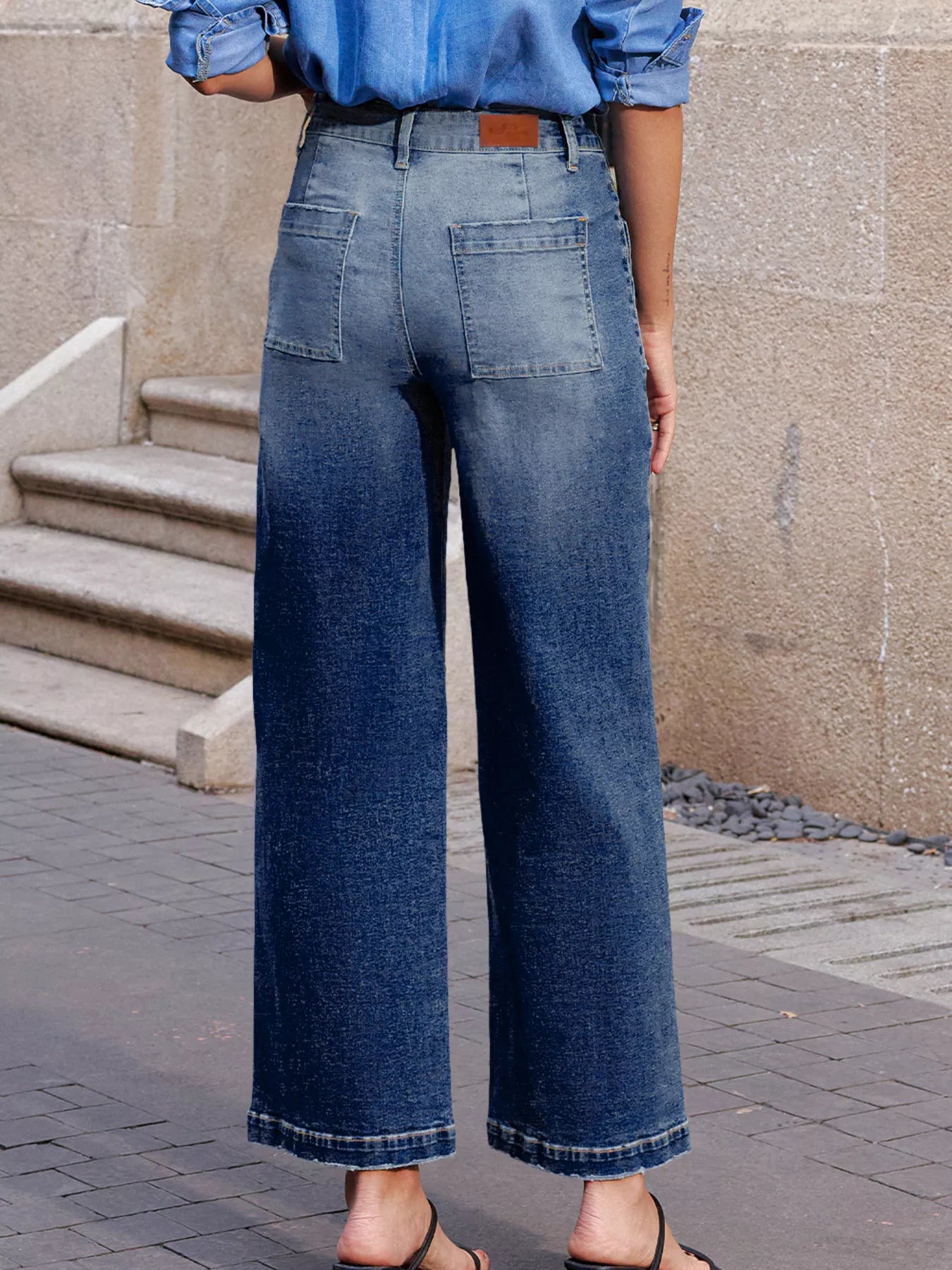 Person wearing blue jeans standing on a sidewalk with a neutral wall background