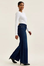 Woman wearing a white long-sleeve top and blue flared jeans on a beige background